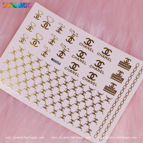 A luxury style water decal sheet, labeled as "WG6056" from Dynamic Nail Supply, is showcased on a pink, fluffy surface. The decals boast gold designs of interlocking double-C logos, the word CHANEL in diverse sizes, and embellishments such as chains and rectangular tags.
