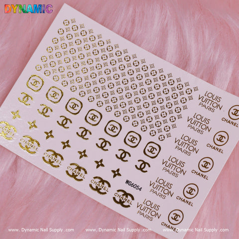 The Luxury Style Water Decal (WG6054) from Dynamic Nail Supply showcases various brand logos such as circular, geometric, and letter designs in a sheet of gold nail art stickers. Elegantly arranged on a soft pink textured background, these decals are ideal for achieving a chic manicure.