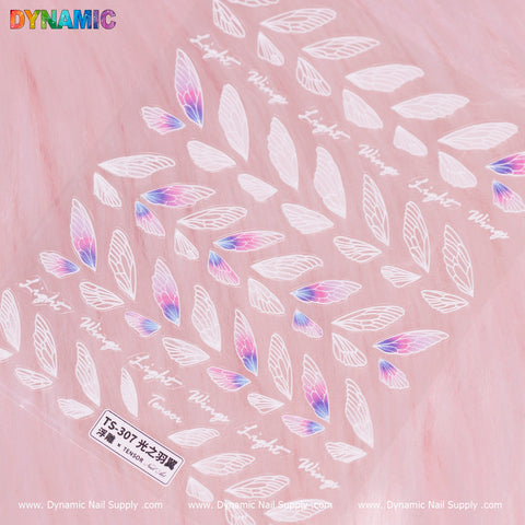 A close-up view of a pink surface highlighting the Dynamic Nail Supply's Wings Sticker (TS - 307), embellished with white and colorful butterfly or fairy wing decals. The design showcases intricate patterns, with wings accented in pink and blue gradients, accompanied by text in various languages among the delicate designs.