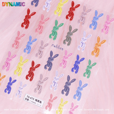 A sheet of Dynamic Nail Supply's Colorful Bunny Stickers (TS-675) showcases vibrant bunnies on a pink background. Arranged in rows, these stickers are available in red, pink, blue, green, and purple hues. Each bunny features the word "rabbit" printed underneath it, with branding text displayed at the top and bottom of the sheet.