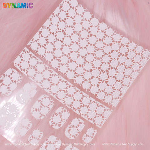 A sheet of White Blossoms Sticker (KX152) from Dynamic Nail Supply is displayed on a pink, furry background. These intricate floral lace nail stickers come in various sizes and designs, ideal for nail art. The word DYNAMIC is prominently showcased in colorful letters at the top left corner.