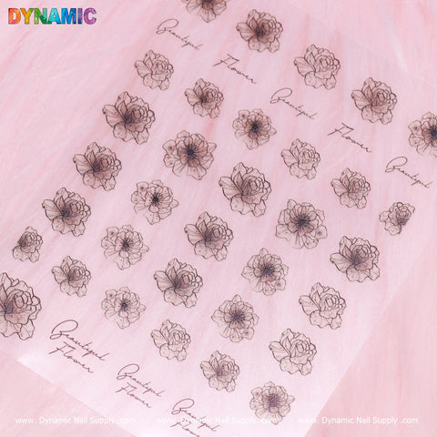 A sheet features the Crystal-black Flower Sticker by Dynamic Nail Supply, set against a soft pink background. The intricate flower designs have delicate black outlines and are interspersed with cursive text that reads "grateful" and "flower." These evenly spaced drawings enhance the sheet with an elegant touch.