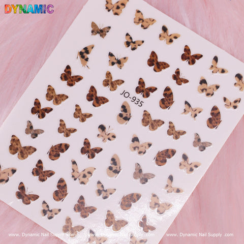 The Pattern Butterfly Sticker (JO-935) features an assortment of brown and black leopard print designs in various sizes, displayed against a soft pink, furry background. The words "Dynamic Nail Supply" are repeatedly printed across the bottom edge of the image.