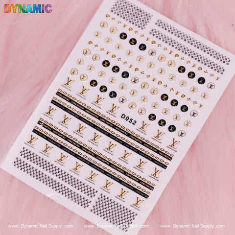 A sheet of the Luxury Nail Art Sticker (D052) by Dynamic Nail Supply is set against a pink background, featuring gold and black designs with logos, patterns, and symbols. The word "DYNAMIC" shines in colorful letters at the top, with the website www.DynamicNailSupply.com elegantly displayed at the bottom.