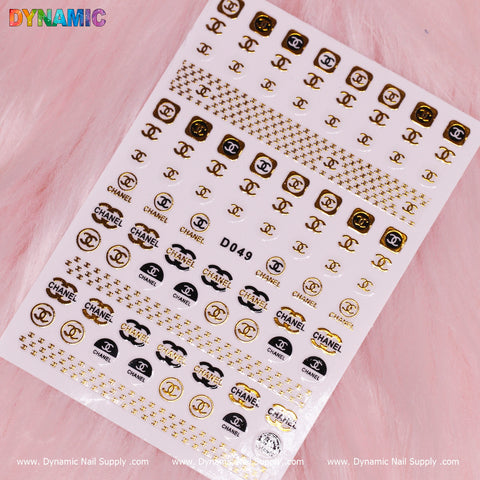 A set of the Luxury Nail Art Stickers (D049) from Dynamic Nail Supply displays brand-inspired logos in stylish black and gold. These stickers are neatly arranged on a white sheet labeled D049, presented against a backdrop of soft pink fur for an elegant touch to any manicure.