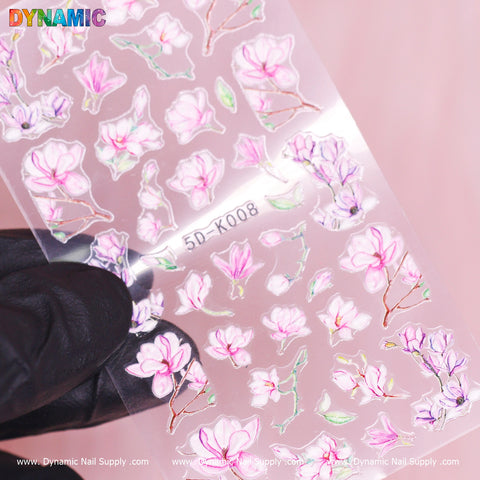 A gloved hand holds a transparent Floral Sticker (5D-K008) sheet from Dynamic Nail Supply, featuring pink and white blossoms with green leaves. Set against a pink background, the sheet is topped with the vibrant DYNAMIC logo, while branding details are visible at the bottom.