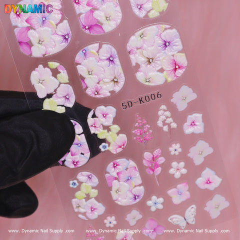 A close-up of the Dynamic Nail Supply Floral Sticker (5D-K006) reveals detailed designs featuring pink, purple, and white flowers highlighted by lush green accents and butterflies. Displayed against a pink background while being held by a hand in a black glove, this artistic piece adds a touch of elegance to any manicure.