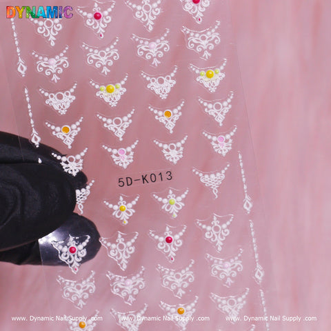 A close-up shot features a nail art sticker sheet displaying detailed white paisley patterns adorned with vibrant gem accents in pink, yellow, and clear hues. These Design Nail Sticker (5D-K013) sheets are transparent and held by a gloved hand, with the Dynamic Nail Supply branding elegantly displayed at the top and bottom.