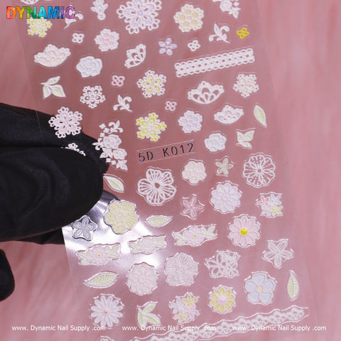 A gloved hand presents a transparent sheet of Floral Sticker (5D-K012) from Dynamic Nail Supply, featuring exquisite designs such as flowers, leaves, and lace patterns. With yellow and green accents, this sheet is ideal for enhancing your nail art experience.