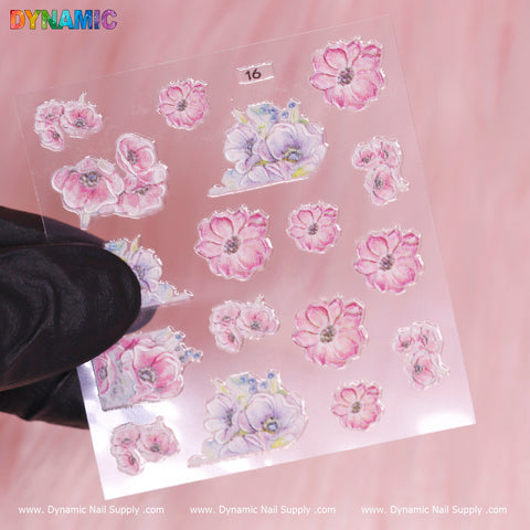 A hand in a black glove holds a transparent sheet of the Flower Sticker (16) from Dynamic Nail Supply, featuring pink and lavender floral designs. The pastel pink background beautifully complements the delicate watercolor style of small clusters and individual flowers, with "Dynamic Nail Supply" elegantly printed along the bottom edge to enhance its charm.