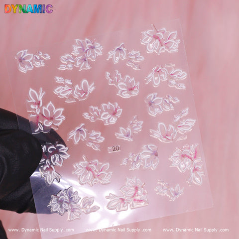 A close-up of a hand wearing a black glove holding a transparent sheet featuring white and pink Flower Sticker (20) for nails. The soft pink background accentuates the vibrant presence of Dynamic Nail Supply at the top, with www.DynamicNailSupply.com repeated along the bottom.
