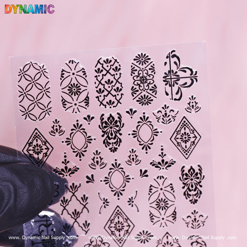 A gloved hand displays a transparent sheet featuring the intricate black "Pattern Nail Sticker (R349)" collection by Dynamic Nail Supply. The stencils showcase floral, geometric, and ornamental designs in shapes such as ovals, diamonds, and semi-circles. Set against a soft pink background, this elegant collection perfectly captures the brand's style.