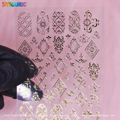 A hand displays a transparent sheet of the Pattern Nail Sticker (R349) - Gold by Dynamic Nail Supply, showcasing geometric shapes, floral patterns, and intricate motifs. The pink background highlights the vibrant multicolored letters in the top left corner. Website addresses are subtly placed at the bottom, with a price tag of R349 discreetly noted.