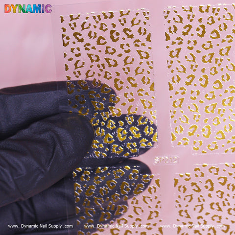 A gloved hand holds a sheet of Animal Pattern Nail Sticker (SP212) featuring a gold leopard print design. The pink background accentuates the elegant pattern, and the brand name Dynamic Nail Supply is prominently displayed at the bottom of the image.