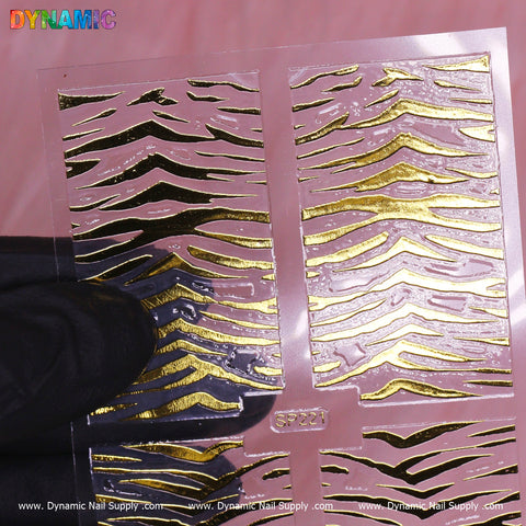 The Animal Pattern Nail Sticker (SP221) by Dynamic Nail Supply showcases gold and black wave designs on a transparent strip. Displayed in a gloved hand against a soft pink fabric background, the nail sticker offers a striking contrast. The elegant branding along the bottom edge enhances its unique design.