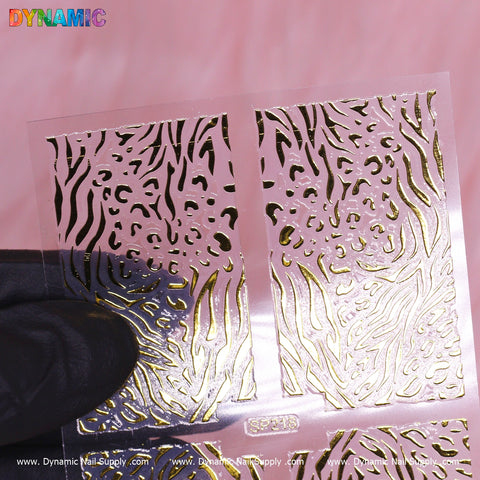 A transparent stencil for nails, known as the Animal Pattern Nail Sticker (SP218), displays intricate animal designs while being held by a person wearing a black glove. The gold zebra and leopard patterns exude elegance. The image is branded with "Dynamic Nail Supply" in colorful letters at the top left corner and includes a website at the bottom.