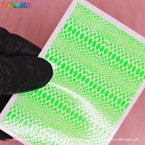A person wearing a black glove holds a rectangular sheet from Dynamic Nail Supply, showcasing the Snake Skin Nail Water Decal in Neon Green. The design is made up of irregular, scalelike shapes that resemble snake skin. The glossy sheet reflects light and prominently features the word DYNAMIC in colorful, bold letters at the top left corner.
