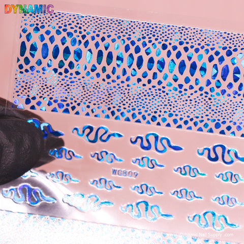 A gloved hand holds the Snake Skin Nail Water Decal (WG809) - Blue Holographic from Dynamic Nail Supply, displayed on a pink background. This decal features wavy and oval patterns, with the upper half highlighting smaller, denser designs and the lower half showcasing larger wavy shapes. The text "DYNAMIC" and "www.DynamicNailSupply.com" is also visible.