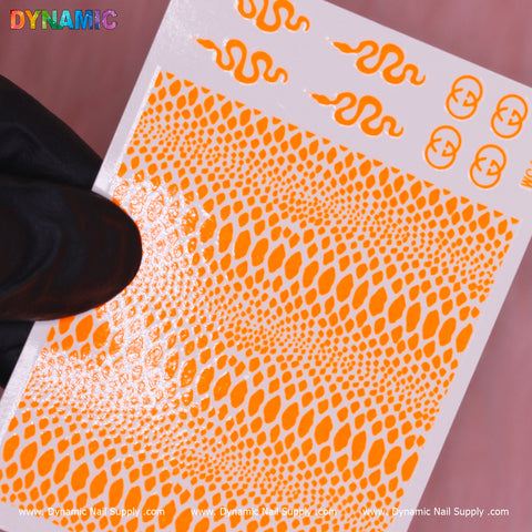 A hand in a black glove displays the Snake Skin Nail Water Decal - Neon Orange from Dynamic Nail Supply against a pink backdrop. The sheet showcases patterns that vary from snake skin textures to serpent shapes, with the "DYNAMIC" logo prominently featured in colorful letters, ideal for those seeking bold, water decal designs.