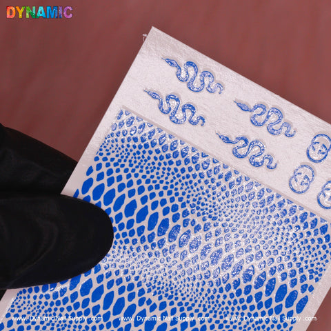 A hand adorned with a black glove displays a sheet of Snake Skin Nail Water Decal - Neon Blue from Dynamic Nail Supply. The decal features an intricate neon blue pattern that resembles snake scales, accompanied by small blue snake designs, against a muted pink background highlighted by the brand name DYNAMIC at the top in a multicolored design.