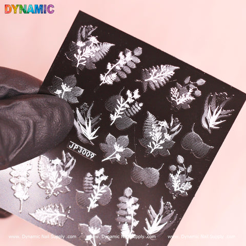 A gloved hand holds a Black and White Plant Sticker (JP3009) by Dynamic Nail Supply against a pale pink background. The sticker features intricate botanical designs, showcasing white patterns of ferns and assorted foliage.