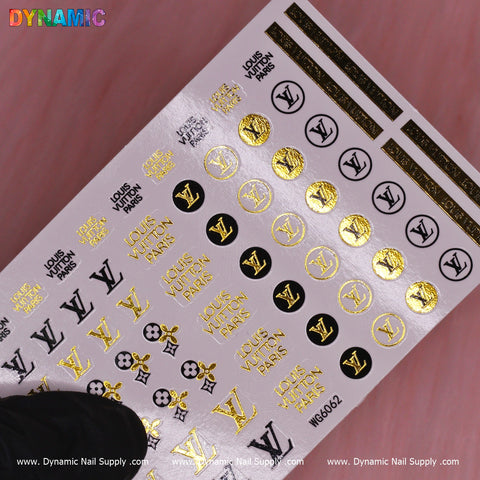 The Luxury Style Water Decal (WG6062) from Dynamic Nail Supply features a collection of brand logos and phrases, predominantly in gold and black, with text displaying LOUIS VUITTON PARIS next to the LV logo. These decals are meticulously arranged in rows against a pink background, offering an elegant presentation with diverse designs.