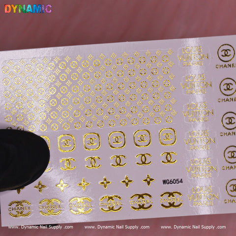 A sheet of Luxury Style Water Decal (WG6054) by Dynamic Nail Supply features opulent brand designs, including interlocking C symbols and Louis Vuitton Paris text. It is placed on a pink surface, with a black object partly visible in the bottom left corner.