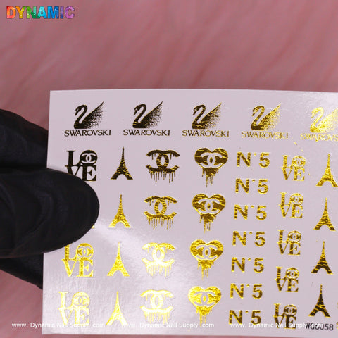 A hand wearing a black glove gently holds a sheet of the Luxury Style Water Decal (WG6058) - Gold by Dynamic Nail Supply, set against a pink background. The gold decals, with designs including the Eiffel Tower, stylized LOVE text, SWAROVSKI's swan logo, and elegantly adorned N° 5 letters, exude luxury.