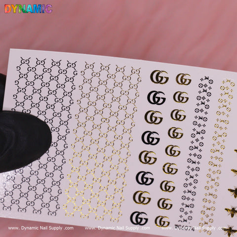 The Luxury Style Water Decal (WG6074) from Dynamic Nail Supply boasts a pattern of interlocking letter designs in gold and black. On the left side, intricate small designs are displayed, while the right features larger bold symbols. A black-gloved hand holds this sheet against a soft pink background.