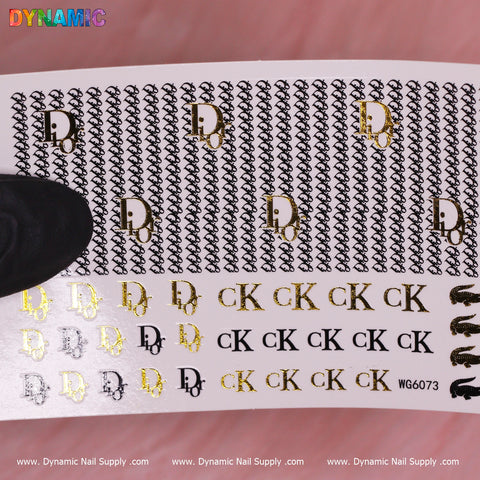 A close-up of a sheet of Luxury Style Water Decal (WG6073) against a light pink background features black and gold monograms, including stylized letters D and CK. A gloved hand holds the sheet, with branding at the bottom reading Dynamic Nail Supply.