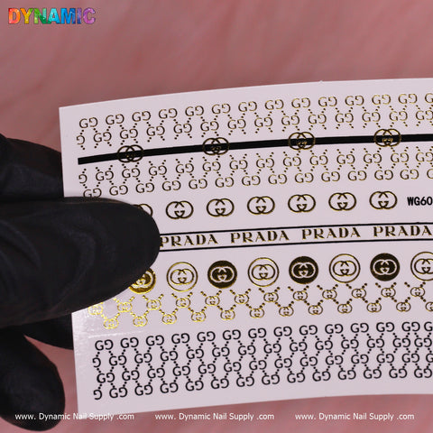 A gloved hand holds a sheet of the Luxury Style Water Decal (WG6069) from Dynamic Nail Supply, featuring intricate metallic gold and black patterns with G G motifs and the word PRADA. Set against a soft pink background, this elegant design exemplifies luxury.