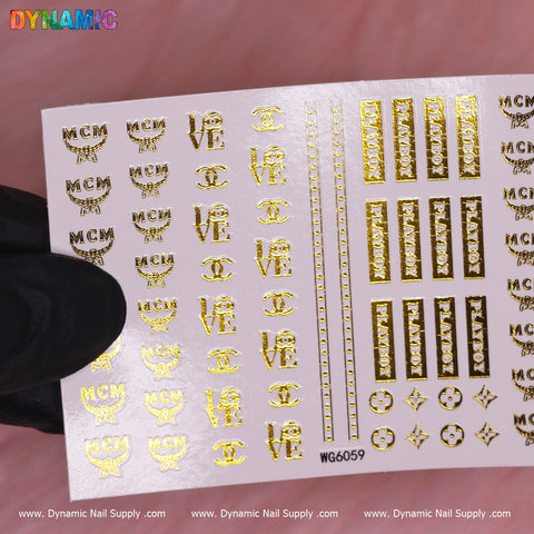 A hand in a black glove holds a sheet of Luxury Style Water Decal (WG6059) - Gold, showcasing an array of luxurious nail art stickers. These water decals include various designs such as MCM, LOVE, and decorative patterns like stripes, stars, and floral motifs. The sheet is branded with Dynamic Nail Supply at the bottom.