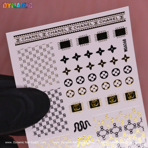 A black-gloved hand holds a sheet of Luxury Style Water Decal (WG6068) nail art stickers. The designs feature iconic logos and patterns in gold, black, and white. Set against a blurred pink backdrop, this sophisticated collection is curated by Dynamic Nail Supply.