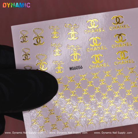 A gloved hand holds a sheet of the Luxury Style Water Decal (WG6056) by Dynamic Nail Supply, showcasing an elegant design with gold nail stickers. Displayed against a soft pink background, this sheet exudes sophistication and features the iconic interlocking CC logo and the word CHANEL.