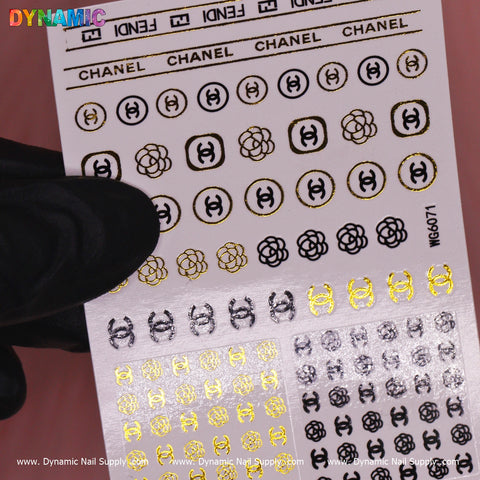 A gloved hand holds a Luxury Style Water Decal (WG6071) from Dynamic Nail Supply, featuring gold, black, and silver nail art stickers adorned with logos, circles, and floral motifs. The branding at the top elevates these designs into exquisite decals for your nails.