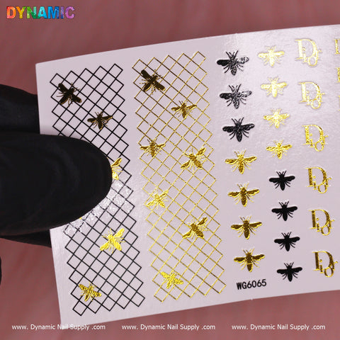 A gloved hand elegantly holds the Luxury Style Water Decal (WG6065) from Dynamic Nail Supply against a soft pink background. This exquisite sheet of nail art stickers showcases golden bees, crisscross patterns, and the letter D in black and gold.