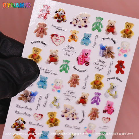 A sheet of adorable Cute Teddy Bear Stickers (R022) from Dynamic Nail Supply features bears in colorful, diverse outfits. Some stickers include phrases like "Classic Teddy" and "Happy Valentine's Day." The sheet is displayed in a gloved hand, highlighting the Dynamic Nail Supply branding.
