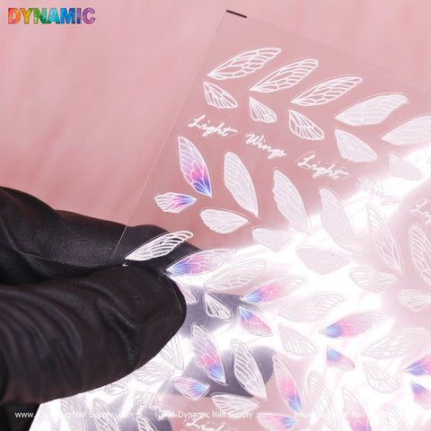 A gloved hand holds a Wings Sticker sheet (product TS - 307) from Dynamic Nail Supply, showcasing iridescent wing-shaped nail decals. These decals exhibit rainbow hues against a shiny silver background, with the brand name "Dynamic" appearing in colorful letters at the top. The sheet features repeated patterns that resemble delicate wings.