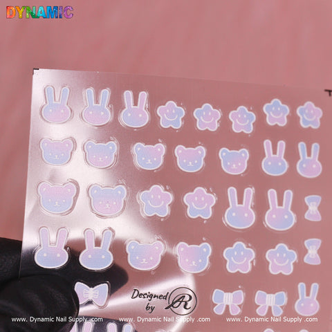 A sheet of vibrant nail art decals from Dynamic Nail Supply features charming animal designs, including the Gradient Bear and Rabbit Sticker. The playful assortment includes smiling pink and blue bunnies, bears, and cats. The soft pink background is headlined with "DYNAMIC," as a black-gloved hand displays the sheet.