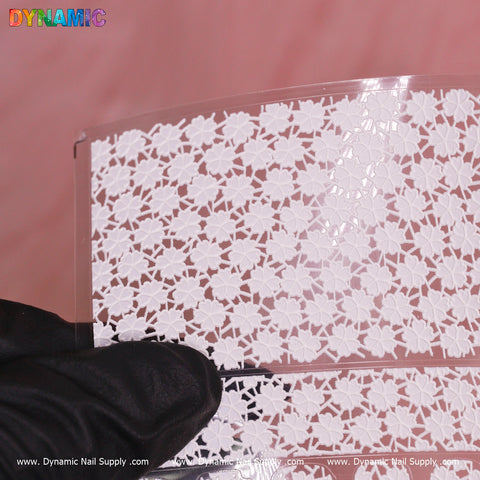 A close-up image of a transparent sheet featuring delicate white floral lace patterns, reminiscent of the White Blossoms Sticker (KX152), held by a gloved hand. The design includes intricate flower and leaf motifs against a soft pink background, with the brand name "Dynamic Nail Supply" repeated along the bottom edge.