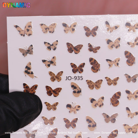 A sheet of the Pattern Butterfly Sticker (JO-935) from Dynamic Nail Supply features an array of sizes and shapes with authentic brown and black leopard spots. These stickers capture the untamed charm of a leopard's pattern and are tastefully displayed by a gloved hand in front of a backdrop adorned with Dynamic Nail Supply logos.