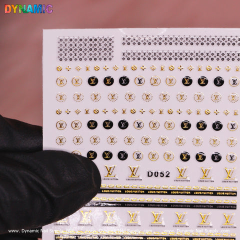 A gloved hand holds a luxurious sheet of the Dynamic Nail Supply Luxury Nail Art Sticker (D052), featuring black and gold designs with logos, stars, and stripes. Set against a soft pink background, the multicolored "DYNAMIC" text in the top left corner stands out prominently.