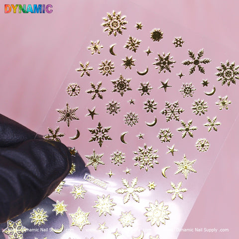 A gloved hand holds a transparent sheet featuring the Christmas Nail Art Sticker - DP2022 from Dynamic Nail Supply, adorned with gold snowflake, star, and crescent moon designs. The bottom displays the label "DP2022," while "DYNAMIC" is printed in multicolored letters at the top against a soft pink background.