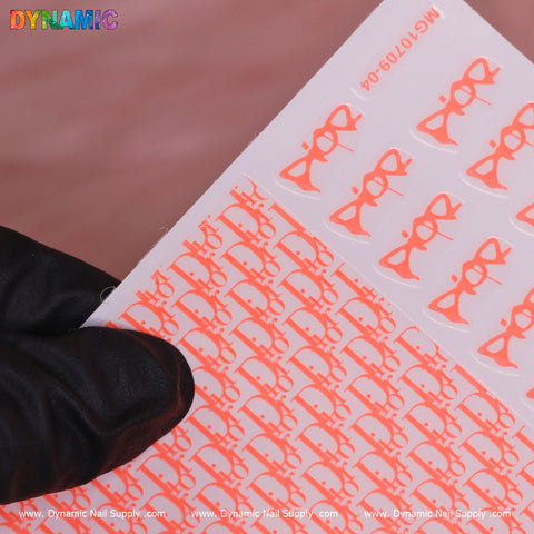 A gloved hand holds the Luxury Water Decal in Neon Red, which features the word Dior in elegant cursive. Set against a soft pink background, Dynamic Nail Supply's branding is prominently displayed at both the top left and bottom border of this stylish nail art accessory.