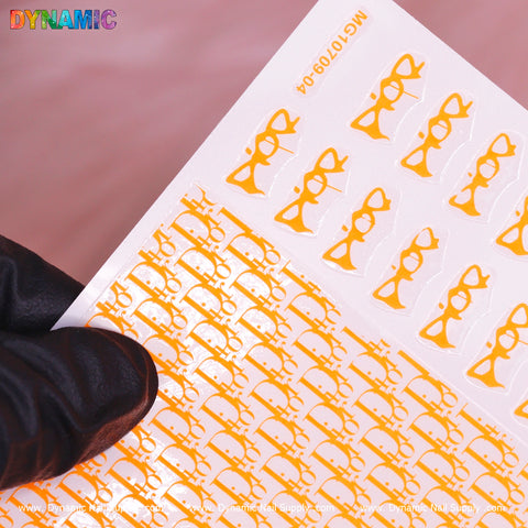 A gloved hand holds a sheet of the Luxury Water Decal (Neon Orange) from Dynamic Nail Supply, showcasing stylish designs featuring neon orange cat-eye glasses. The sheet is highlighted against a soft pink background, with "DYNAMIC" and "www.DynamicNailSupply.com" elegantly adorning the top and bottom edges.