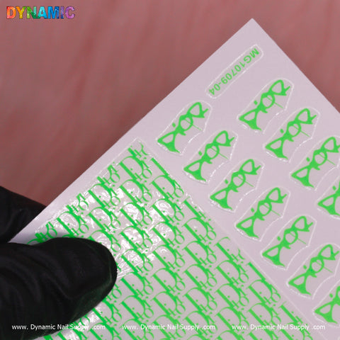 A gloved hand holds a sheet of the Luxury Water Decal (Neon Green) from Dynamic Nail Supply, featuring eyeglasses against a vibrant pink background. The decals are neatly arranged in rows, with www.DynamicNailSupply.com elegantly printed along the bottom edge, highlighting their exclusive allure.