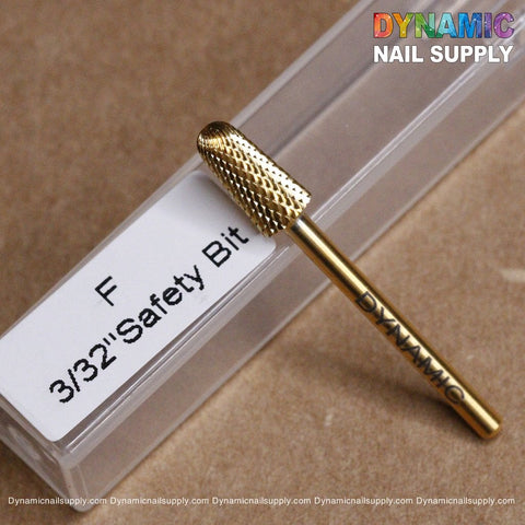 A gold-colored F Safety Bit is placed on a brown surface. The textured bit sits beside a clear rectangular case labeled "F 3/32” Safety Bit." In the background, the vibrant watermark of Dynamic Nail Supply decorates the corners, showcasing its precision like a genuine Dynamic Drill Bit.
