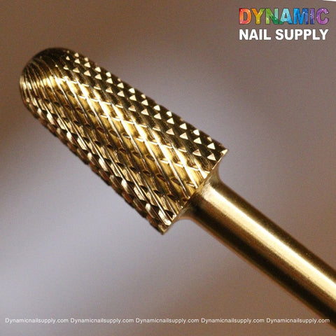 Close-up of a gold-colored, cylindrical F Safety Bit - Dynamic Drill Bit showcasing sharp, diamond-shaped ridges. The logo for Dynamic Nail Supply appears in colorful letters in the top right corner, with a partially visible website URL repeated in small text at the bottom.