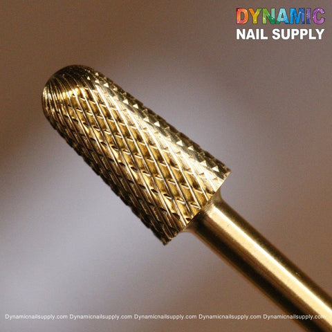 This is a close-up of a gold-colored, textured F Safety Bit - Dynamic Drill Bit with a cylindrical shape and diamond-patterned surface designed for nail care. The softly blurred background enhances its allure. Featuring the brand name "Dynamic Nail Supply" in colorful letters, with the website subtly repeated at the bottom.