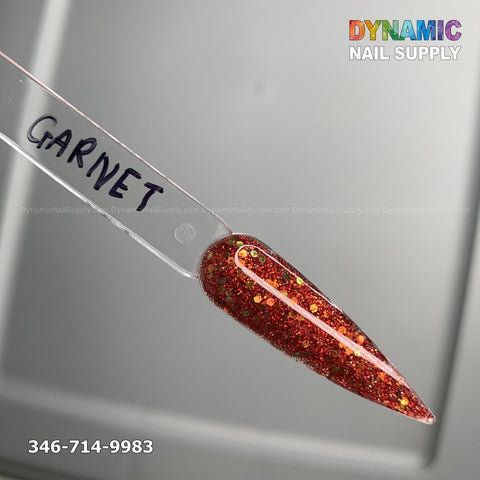 A long, pointed nail swatch labeled Garnet - Acrylic Glitter Powder displays a sparkling red polish infused with gold and multicolored flecks. This non-toxic formula is designed to ensure long-lasting nails. The background highlights the Dynamic Nail Supply logo in vibrant lettering, with the phone number 346-714-9983 located at the bottom left corner.